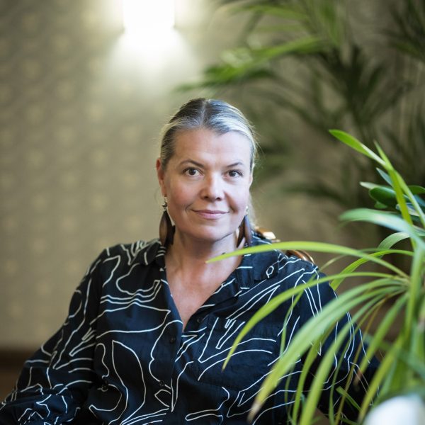 Keski-ikäinen nainen istuu tapetoidussa huoneessa ja katsoo kameraan ja hymyilee